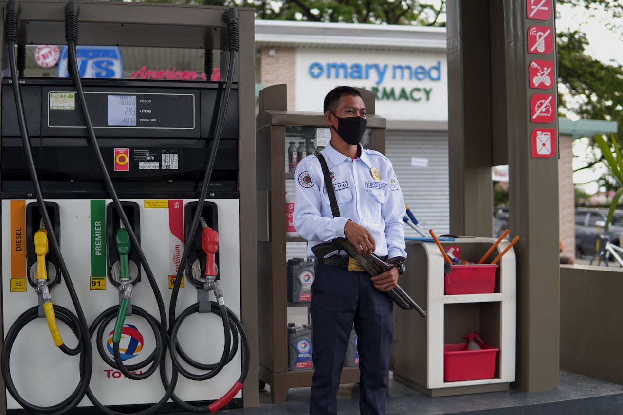 A uniformed security guard wearing a face mask stands vigilant at a gasoline station with fuel pumps.